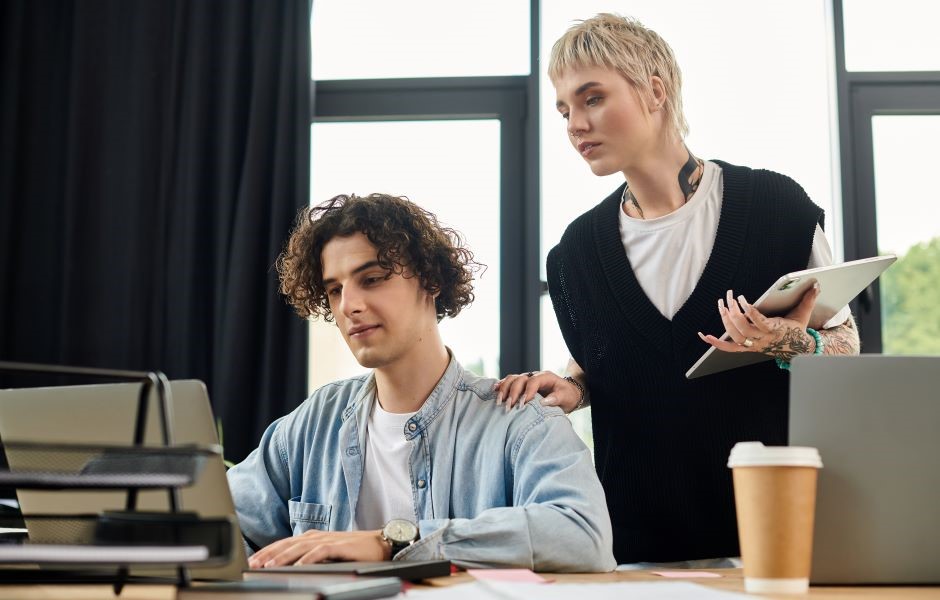 Imagem de uma jovem profissional ajudando um jovem a trabalhar no escritório, com ambos usando laptops e materiais de escritório ao redor, destacando colaboração e produtividade.