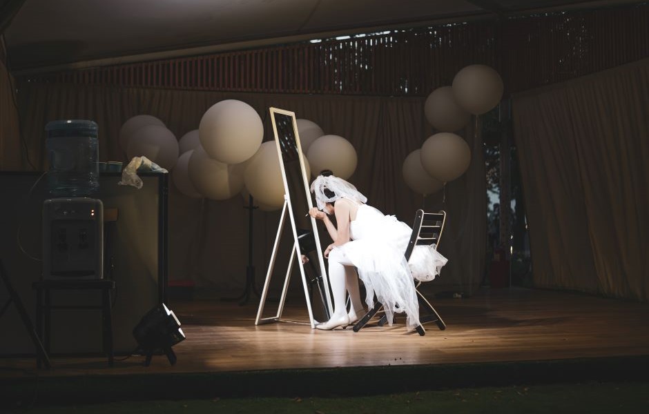 No palco, uma atriz com vestido branco e maquiagem artística está pintando ou desenhando em um espelho grande, decorada com balões brancos e iluminação suave. Cena de evento artístico ou espetáculo de teatro.