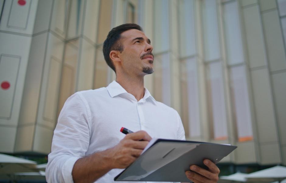 Um homem de camisa branca olha pensativo para cima enquanto segura uma prancheta e uma caneta.