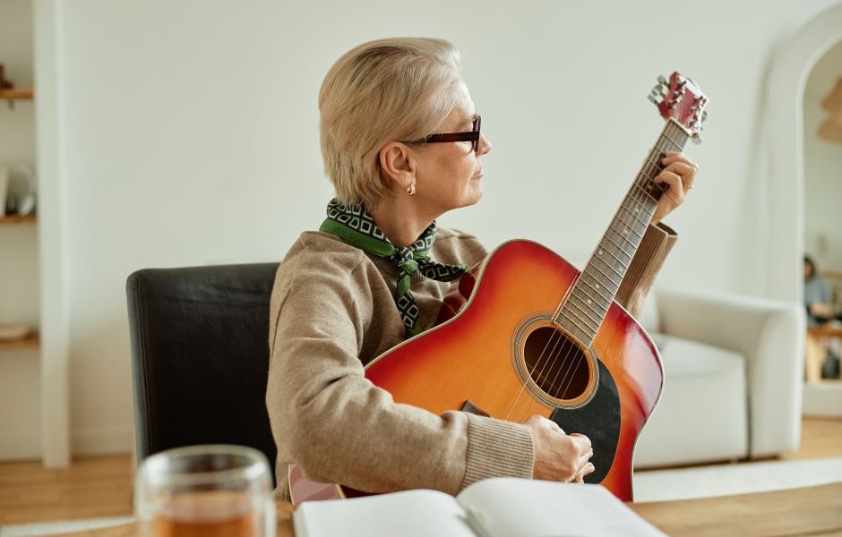 Mulher idosa tocando violão em ambiente aconchegante, vestindo óculos e com cabelo grisalho, criando um momento de música e relaxamento em casa.