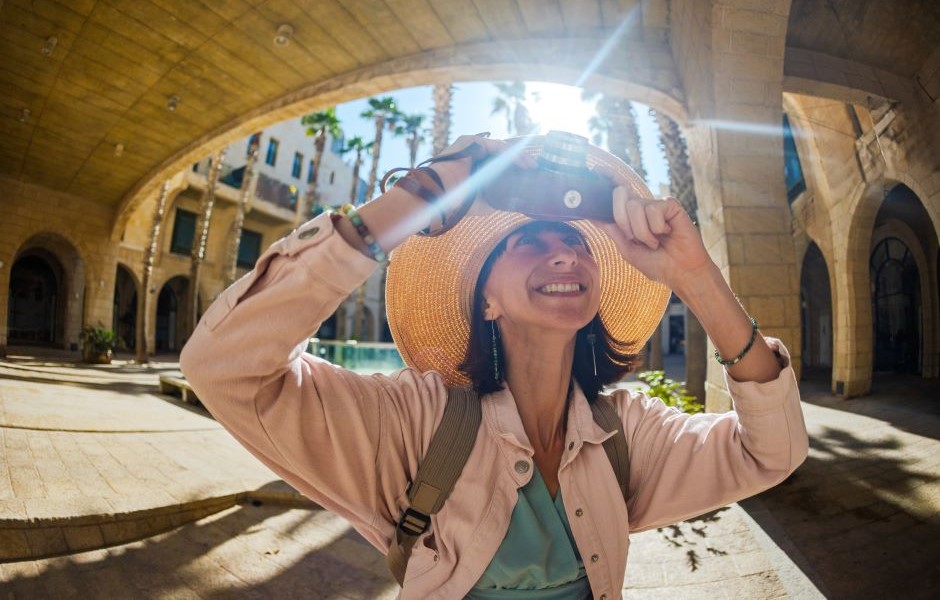 Mulher sorridente com chapéu de palha tomando uma selfie ao ar livre em um ambiente com arquitetura antiga e árvores, aproveitando um dia ensolarado de viagem.