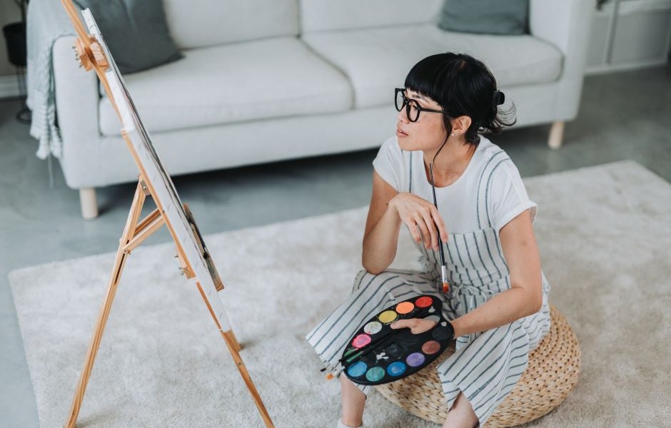 Mulher pintando em casa com um vestido listrado e óculos, sentada em um puff de palha com uma paleta de cores vibrantes ao lado, ao lado de um cavalete com uma tela. O ambiente é acolhedor, com tapete claro e sofá cinza ao fundo.