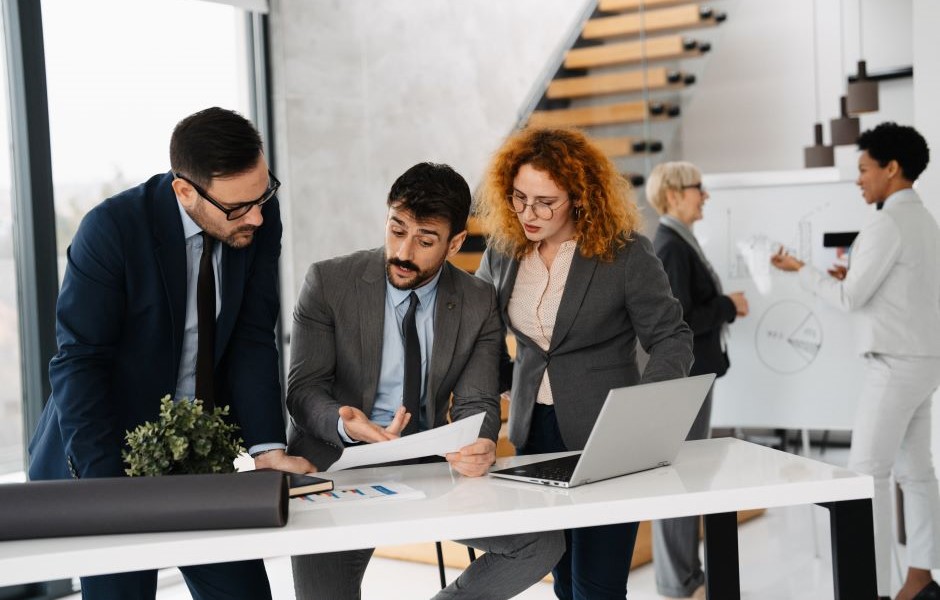 Equipe de profissionais de negócios discutindo estratégias durante uma reunião no escritório, com colegas colaborando ao fundo, transmitindo um ambiente de trabalho dinâmico e profissional.