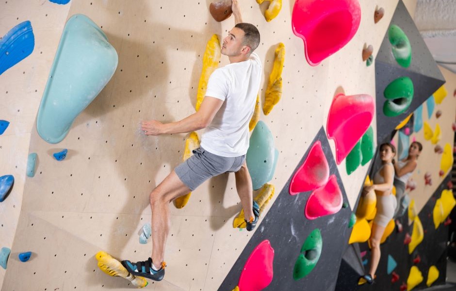 Homem praticando escalada em parede de boulder no ginásio de esportes, com outros praticantes ao fundo, em ambiente interno.