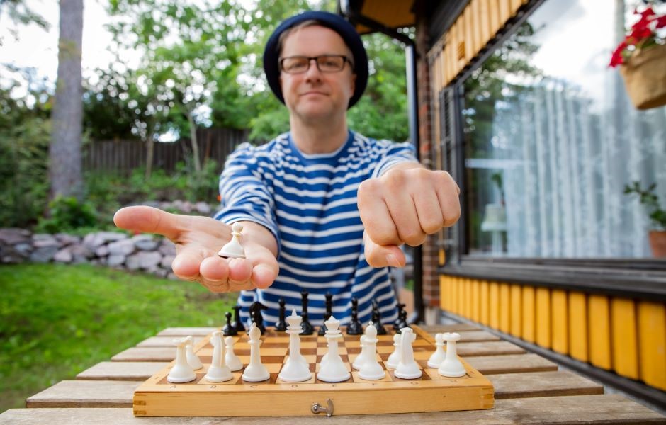 Homem jogando xadrez ao ar livre, vestido com camisa listrada azul e branca, usando óculos e chapéu, com fundo de jardim e casa de campo ao fundo.